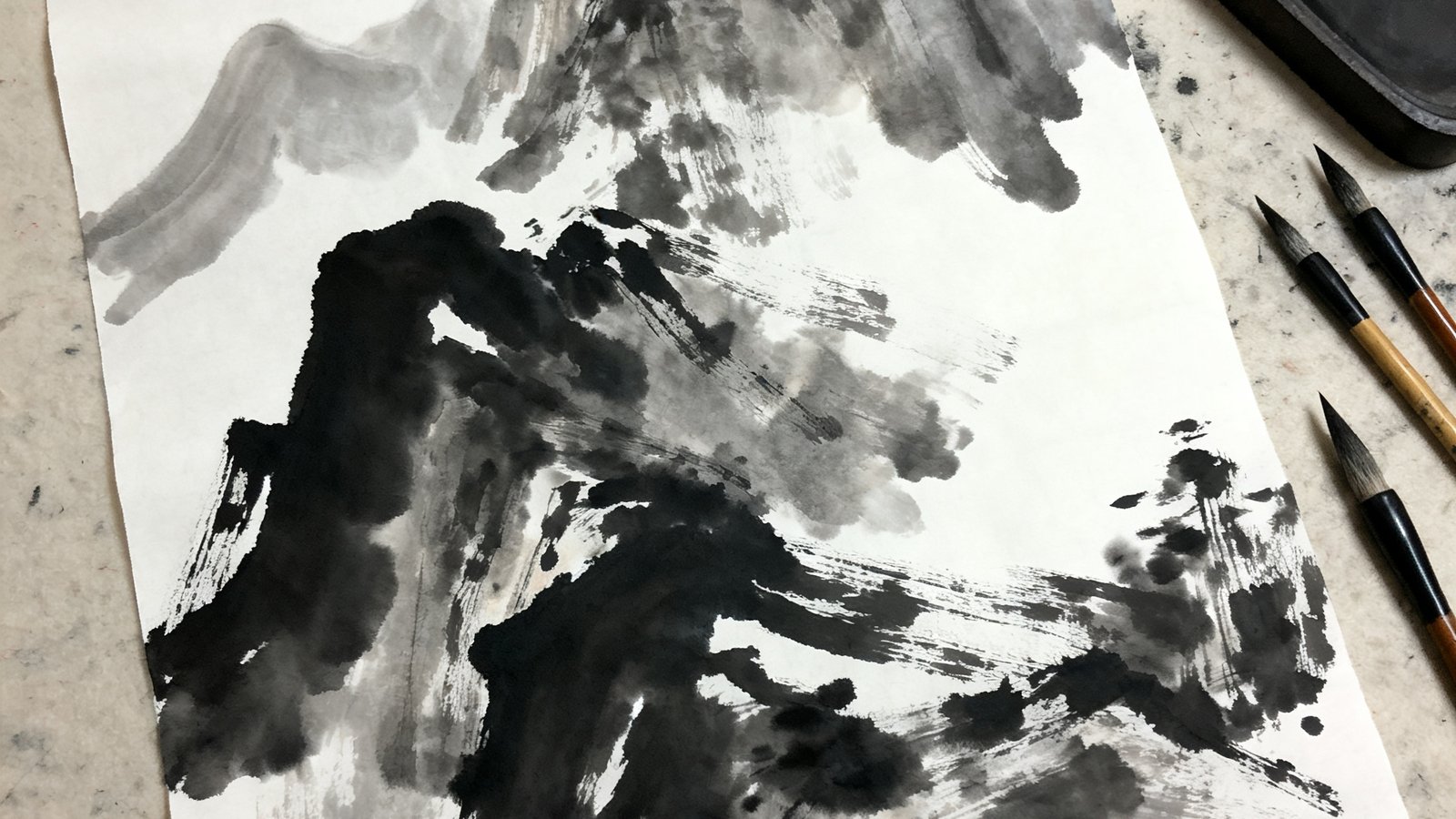 一幅当代水墨作品的特写，墨色在宣纸上晕染出山峦的轮廓，画面留白恰到好处，笔触既有传统功力又充满现代构成感，旁边散落着几支毛笔和一方端砚。